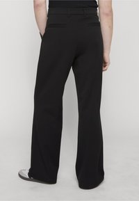 Person wearing black wide-leg pants with back pockets, standing with hands in pockets, showing the rear view from waist to feet.