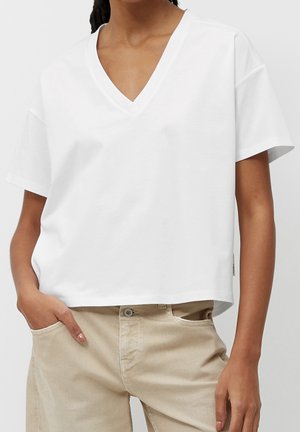 Osoba ubrana w luźną białą bluzkę z krótkim rękawem i dekoltem w kształcie litery V oraz beżowe spodnie, z jedną ręką w kieszeni, widziana od ramion do połowy uda.