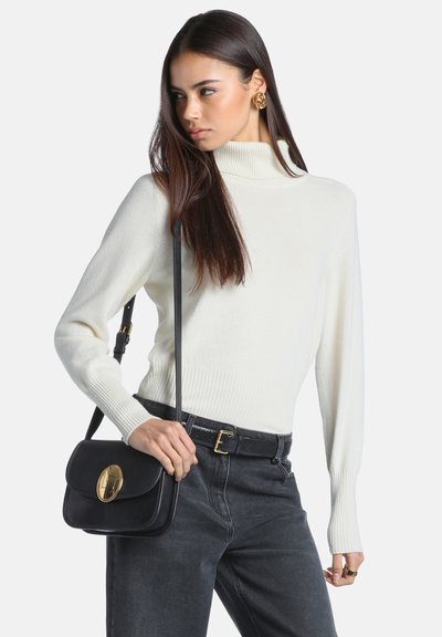 Femme portant un pull à col roulé blanc et un jean noir, tenant un sac à bandoulière noir avec une boucle dorée, regardant sur le côté.