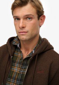Sudadera con cremallera marrón sobre una camisa de franela de cuadros en verde y azul. La sudadera presenta un logo en rojo, con tejido texturizado, de estilo casual.