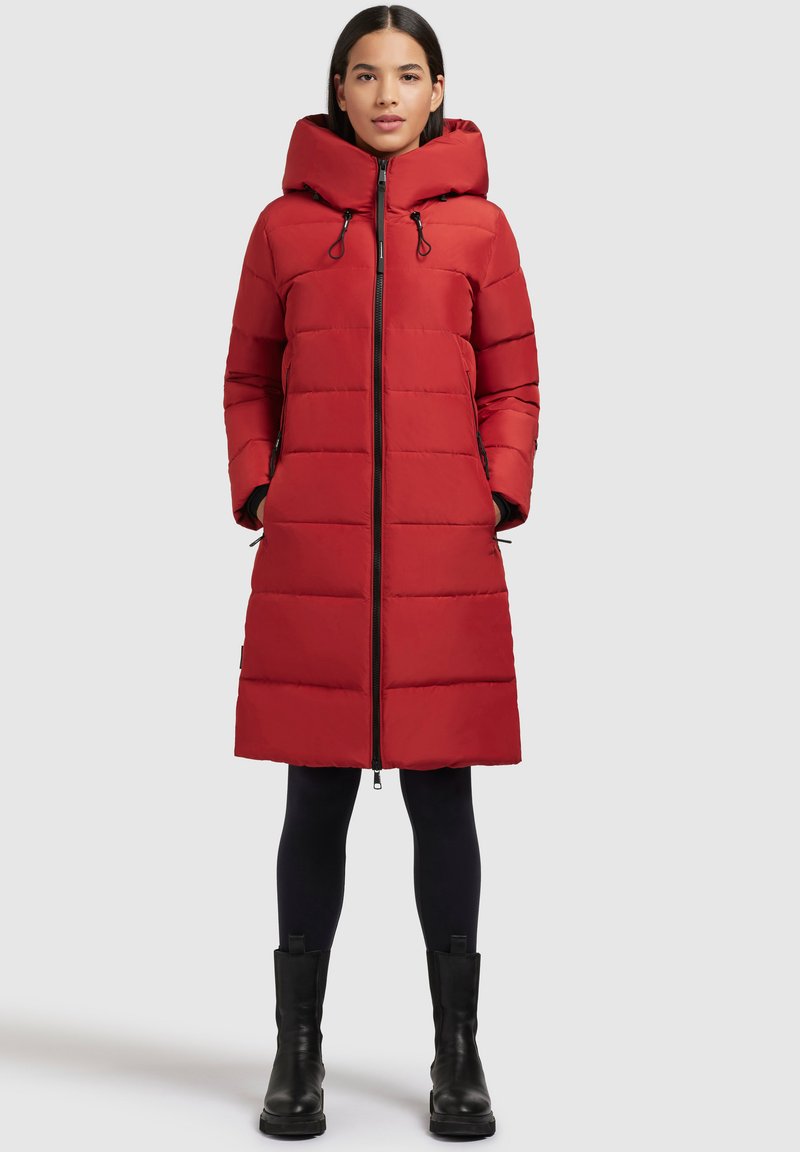 Vrouw in een lange rode gewatteerde pufferjas met capuchon, zwarte leggings en zwarte laarzen, staande met haar handen in de zakken.