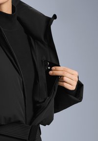 Czarna kurtka z wysokim kołnierzem, wyposażona w kieszeń na piersi zapinaną na zamek. Ręka wkłada smartfon do kieszeni. Widoczna faktura materiału.