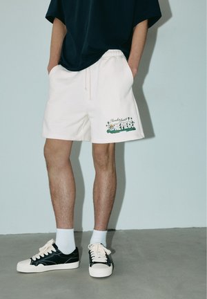 Personne portant un short blanc avec un logo One Piece, des baskets noires et blanches, des chaussettes blanches et un haut sombre, debout les mains dans les poches.