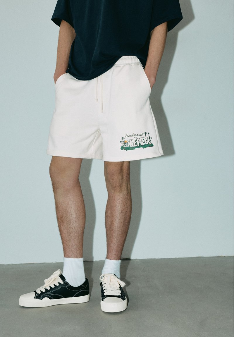 Personne portant un short blanc avec un logo One Piece, des baskets noires et blanches, des chaussettes blanches et un haut sombre, debout les mains dans les poches.