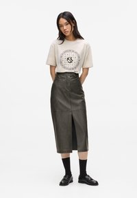 Khaki kožená midi sukňa s predným rozparkom, kombinovaná s béžovým grafickým tričkom a čiernymi ponožkami. Model má na sebe čierne mokasíny.