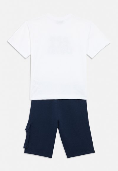 T-shirt blanc à manches courtes associé à un short bleu marine arrivant aux genoux, doté d'une poche cargo latérale avec fermeture à bouton.