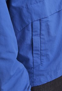Blaue Jacke aus einem glatten, leichten Material mit matter Oberfläche, diagonalen Nahtdetails und einer gefalteten Tasche an der Seite.