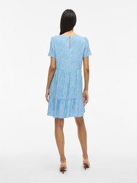 Niebieska sukienka z krótkim rękawem, z drapowanym, warstwowym dołem, zdobiona kropkowanym wzorem i otworem w kształcie łezki z tyłu. Gładka tkanina o lekkiej fakturze.