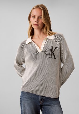 Jeune femme aux cheveux blonds porte un haut rayé noir et blanc à manches longues avec un col blanc et un logo noir "CK", associé à un jean bleu.
