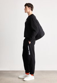 Juodas megztinis ir sportinės kelnės, abi su laisva pritaikymu. Balta "Calvin Klein" logotipas ant kelnių. Baltos sportbačiai ir raudonos kojinės užbaigia aprangą.