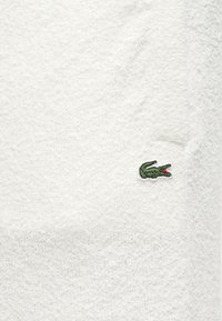 Tissu éponge blanc avec une texture douce, arborant un petit logo de crocodile vert dans le coin inférieur. Design simple et épuré.