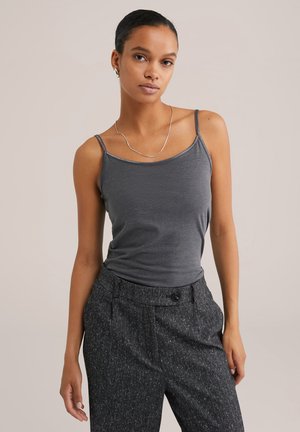 Jeune femme aux cheveux bruns, portant un haut sans manches gris et un pantalon à motifs sombres, se tient devant un fond clair.