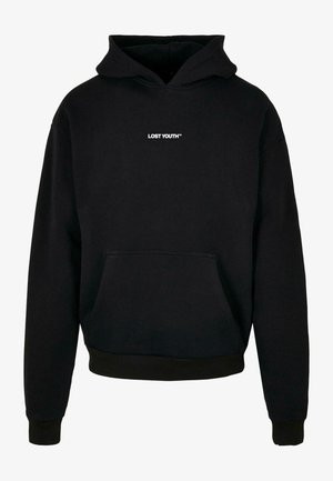 Persona che indossa una felpa con cappuccio nera con la scritta bianca "LOST YOUTH™" sul petto, abbinata a pantaloni grigio chiaro e un tatuaggio sulla mano visibile.