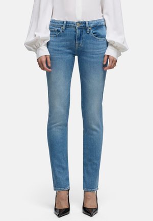 Kvinde iført lyseblå tætsiddende jeans, hvid langærmet bluse med puffede manchetter og sorte spidse høje hæle, stående mod en ensfarvet baggrund.