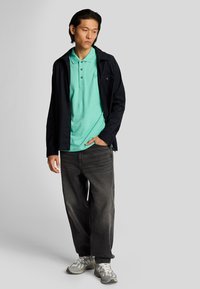Camisa polo verde menta debajo de una chaqueta negra, combinada con unos vaqueros grises holgados y zapatillas grises. El conjunto presenta un look casual y en capas.