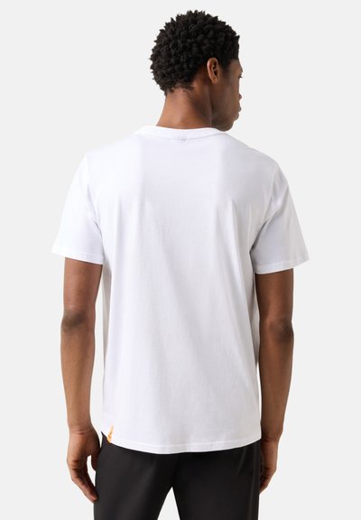 Homme aux cheveux courts et bouclés portant un t-shirt blanc uni à manches courtes et un pantalon noir, vu de dos sur un fond uni.