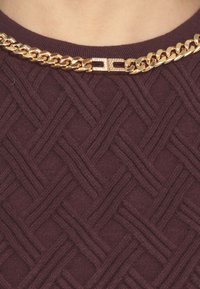 Suéter burdeos con un patrón tejido en relieve. Acompañado de un collar de cadena dorada con un cierre decorativo.