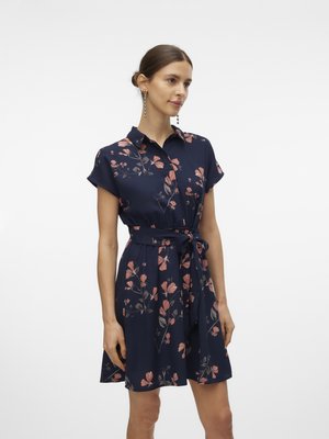 Donna che indossa un vestito camicia a maniche corte blu navy con fantasia floreale e cintura in vita, in piedi davanti a uno sfondo bianco semplice.