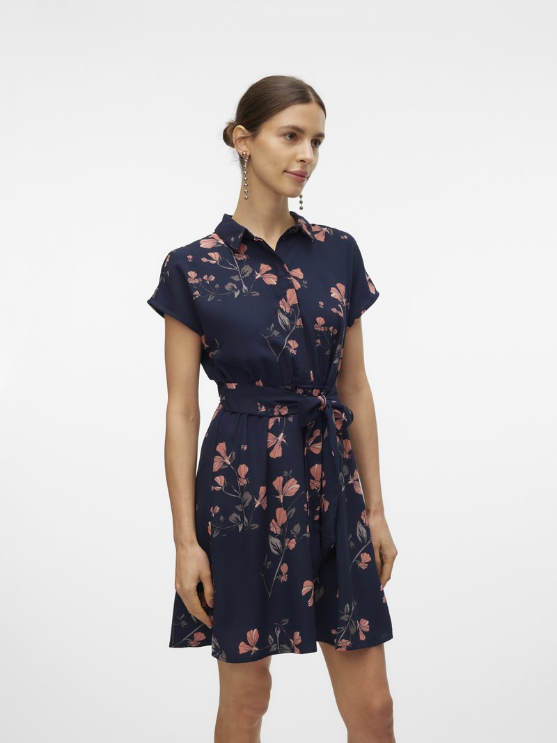 Žena u tamnoplavoj cvjetnoj haljini s kratkim rukavima i remenom u struku, stoji na jednobojnoj bijeloj pozadini.