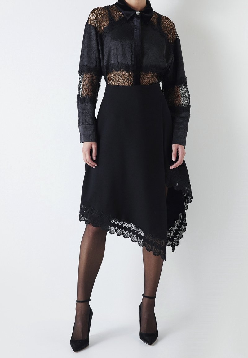 Femme portant un chemisier en dentelle noire à manches longues et une jupe noire asymétrique avec bordure en dentelle, des collants noirs transparents et des talons noirs.