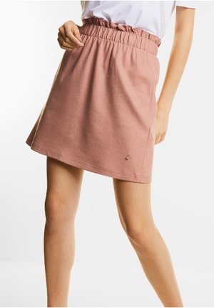 Femme portant une jupe à taille élastique rose poussiére associé à un haut blanc à manches courtes, debout devant un fond blanc uni.