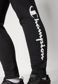 Svarte joggebukser laget av bomullsblanding, med en hvit "Champion"-logo trykket vertikalt langs benet. Ribbede mansjetter og matchende svarte joggesko.
