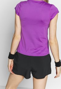 Top deportivo morado de mangas cortas con tejido texturizado, combinado con pantalones cortos negros de corte holgado y dobladillo curvado en la parte inferior. Se puede ver una pulsera negra.