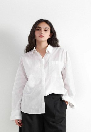 Junge Frau mit langen dunklen Haaren, die ein locker sitzendes weißes, gestreiftes Hemd und schwarze Hosen vor weißem Hintergrund trägt.