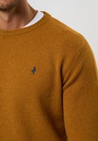 Senfgelbes Strickpullover mit einem gerippten Rundhalsausschnitt, ausgestattet mit einem kleinen dunkelblauen Logo auf der linken Brust; glatte Textur.
