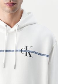 Witte hoodie met trekkoorden, met een zwart "CK" logo en een blauwe Calvin Klein merkstreep over de borst.