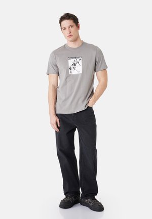 Hombre con el pelo corto y rizado que lleva una camiseta gris con temática de hockey, vaqueros negros y zapatillas negras, de pie con una mano en el bolsillo.