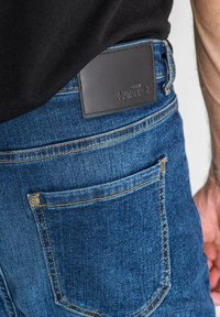 Niebieskie jeansy z denimu z kontrastowym żółtym przeszyciem, wyposażone w tylną kieszeń oraz czarną skórzaną metkę z wytłoczonym napisem "KAVANACH".