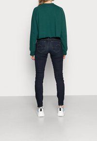 Jeans skinny azul oscuro con cintura alta, acompañados de un sudadera corta verde. Los zapatos son blancos con acentos en azul claro.