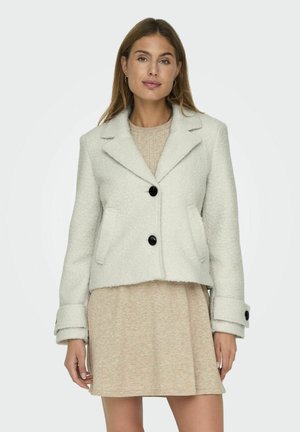 Weiße cropped Jacke mit schwarzen Knöpfen und Taschen, aus strukturiertem Stoff. Über einem beigen Strickkleid getragen.