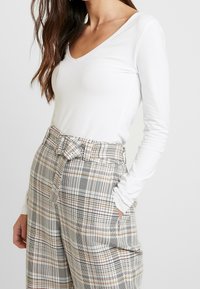 Langärmliges weißes V-Ausschnitt-Shirt verbunden mit hoch taillierten karierten Hosen, die eine Mischung aus schwarzen, braunen und roten Linien sowie einem Gürtel in der Taille aufweisen.