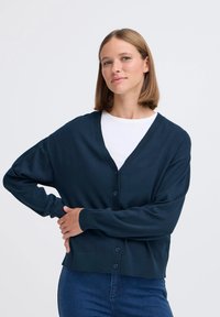 Cardigan blu navy con scollo a V, maniche lunghe e chiusura a bottoni. Texture morbida con dettagli a coste sui polsini e sull'orlo. Indossato sopra una maglietta bianca.