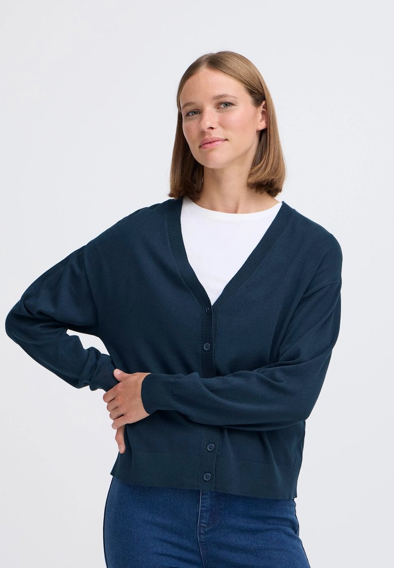Cardigan blu navy con scollo a V, maniche lunghe e chiusura a bottoni. Texture morbida con dettagli a coste sui polsini e sull'orlo. Indossato sopra una maglietta bianca.