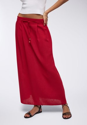 Gonna maxi leggera in lino rosso con vita a cordoncino, caratterizzata da un design fluido e una texture minimale. Abbinata a sandali neri con cinturini.
