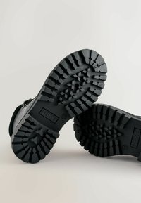 Semelles de bottes en caoutchouc noir avec des motifs de semelle profonds et le nom de la marque « NEXT » embossé. Présente un design robuste pour une bonne traction.
