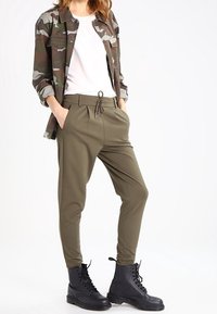 Jacka med camouflage-mönster över en vit skjorta; olivgröna joggerbyxor med elastiskt midjeband och dragsko; svarta kängor.