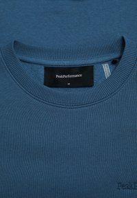 Felpa di cotone blu con colletto a girocollo, polsini e orlo a costine. Presenta un'etichetta PeakPerformance sul colletto e un dettaglio del logo ricamato.
