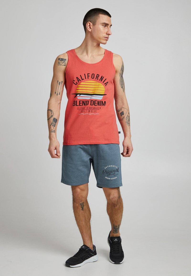 Rød tanktop med California-grafik og gult solmotiv, kombineret med blå shorts med logo. Båret med sorte atletiksko.