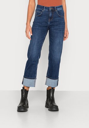 Frau trägt dunkelblaue Jeans mit Umschlag und schwarze klobige Stiefeletten, steht vor einem schlichten hellen Hintergrund.