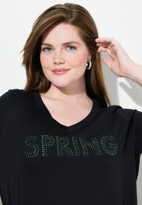 Schwarzes V-Ausschnitt-T-Shirt mit grüsem Strass-Text "SPRING". Weicher Stoff mit lockerer Passform, verziert mit dezenten Glitzerdetails.