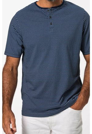 Homme portant un polo manches courtes henley rayé bleu marine et bleu clair avec trois boutons, associé à un pantalon blanc, une main dans la poche.