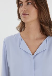 Blusa azzurro chiaro con colletto a punte e chiusura con bottoni. Tessuto liscio con un leggero alone e maniche lunghe. Design minimalista.