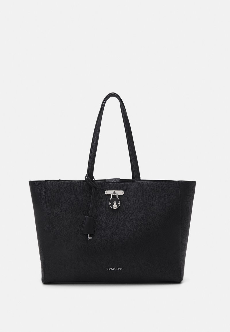 Sac cabas en cuir noir Calvin Klein avec double poignée, détail de cadenas et porte-clés argentés, texture grainée sur fond blanc.