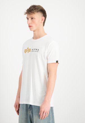 Hvid bomulds t-shirt med et guld- og sort "ALPHA INDUSTRIES" logo, rund hals, korte ærmer og et branding-tryk på ærmet.