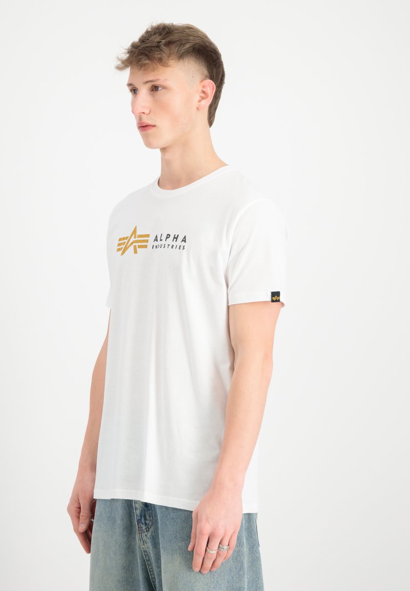 Bílý bavlněný tričko s kultovním černým a zlatým logem "ALPHA INDUSTRIES", kulatý výstřih, krátké rukávy a brandingový detail na rukávu.