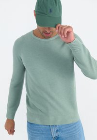 Hellgrüner, gerippter Strickpullover mit Rundhalsausschnitt und langen Ärmeln, kombiniert mit einer passenden grünen Mütze, lässig getragen.